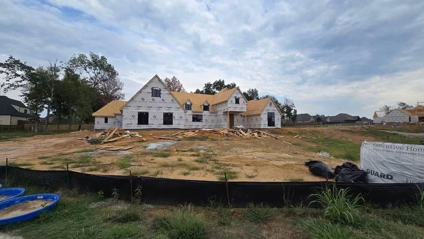 Two-story home under construction, wrapped in white house wrap with roof sheathing installed and framed window openings